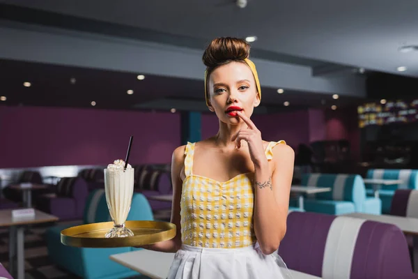 Neugierige Pin-up-Kellnerin mit Tätowiertablett und Milchshake im Café — Stockfoto