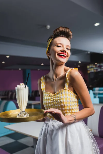 Joyful pin up waitress with tattoo holding tray and tasty milkshake in cafe — Stock Photo