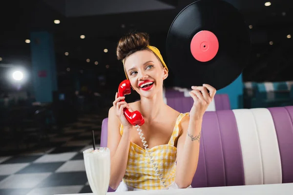 Glückliche Pin-up-Frau mit roten Lippen hält Retro-Schallplatte in der Hand und telefoniert in der Nähe von Milchshake im Café — Stockfoto