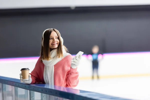 Mujer complacida en orejeras blancas y suéter rosa sosteniendo taza de papel y teléfono celular en pista de hielo - foto de stock