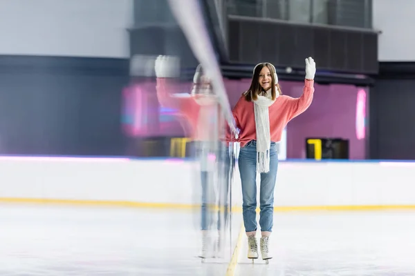 Pleased woman in white ear muffs and pink sweater skating and waving hand on ice rink — Stock Photo