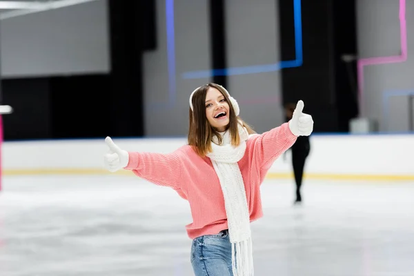 Amazed young woman in pink sweater showing thumbs up on ice rink — Stock Photo