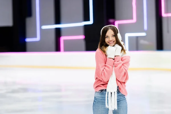 Cheerful young woman in pink sweater looking at camera on ice rink — Stock Photo