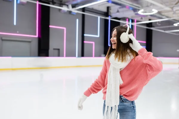 Молода весела жінка в білому шарфі, що регулює вушні муфти і катається на ковзанах — стокове фото