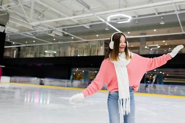 Возбужденная женщина в белых наушниках и розовом свитере катается на коньках с протянутыми руками на катке — стоковое фото