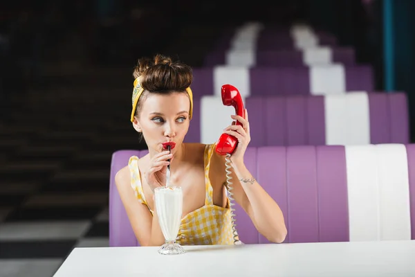 Junge Pin-up-Frau hält rotes Handy in der Hand und trinkt köstlichen Milchshake im Café — Stockfoto