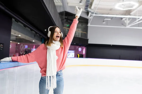 Mujer feliz en orejeras blancas y suéter rosa patinaje con la mano extendida en pista de hielo - foto de stock