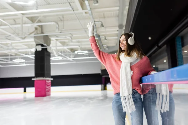 Happy woman in white gloves, ear muffs and pink sweater waving hand on ice rink — Stock Photo