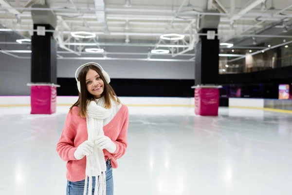 Femme heureuse en gants blancs, cache-oreilles et pull rose souriant sur la patinoire — Photo de stock