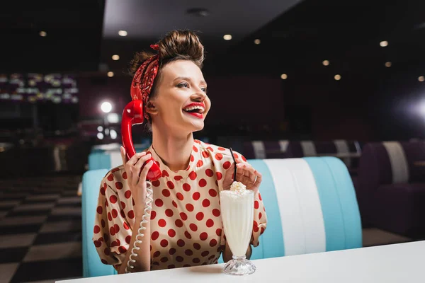 Happy pin up woman with red lips talking on retro telephone near milkshake on table — Stock Photo