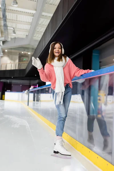 In voller Länge glückliche junge Frau im Winteroutfit, die auf gefrorener Eisbahn Schlittschuh läuft und winkt — Stockfoto