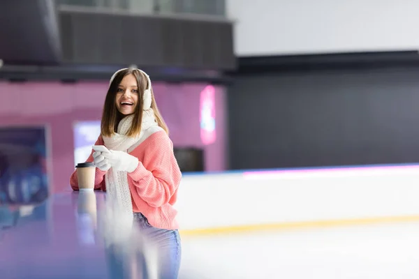 Femme excitée dans des cache-oreilles tenant smartphone près de tasse en papier sur la patinoire — Photo de stock