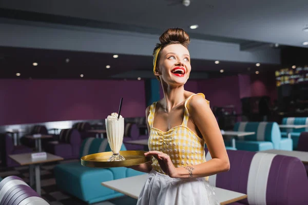 Garçonete pin up alegre com bandeja de tatuagem segurando com milkshake no café — Fotografia de Stock