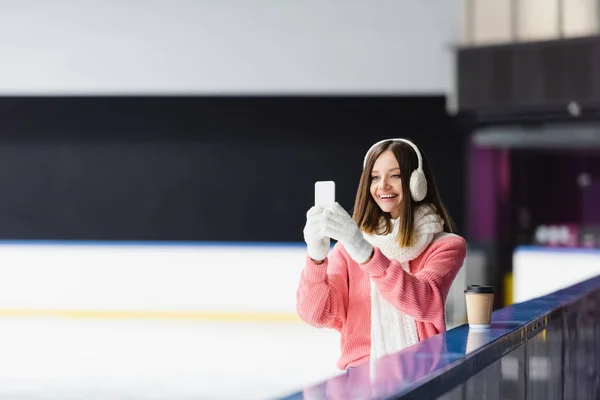 Femme heureuse dans des cache-oreilles et des gants prenant des photos sur la patinoire — Photo de stock