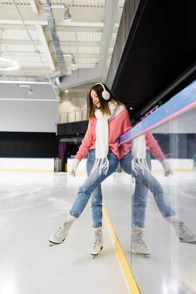 Comprimento total de alegre jovem mulher em roupa de inverno olhando para patins de gelo na pista congelada — Fotografia de Stock