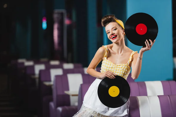 Pin up mujer con labios rojos sosteniendo discos de vinilo retro y sonriendo en la cafetería - foto de stock