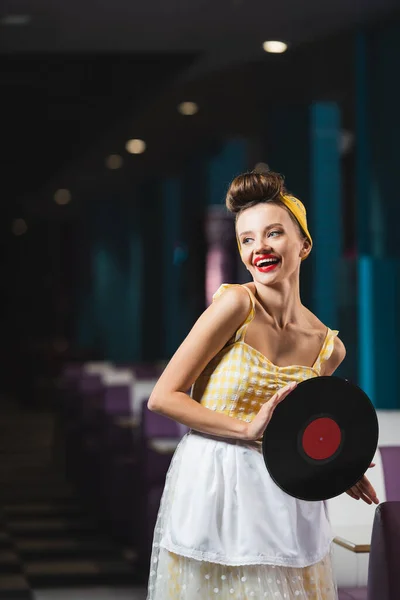 Alegre pin up mujer con labios rojos sosteniendo disco de vinilo retro y sonriendo en la cafetería - foto de stock