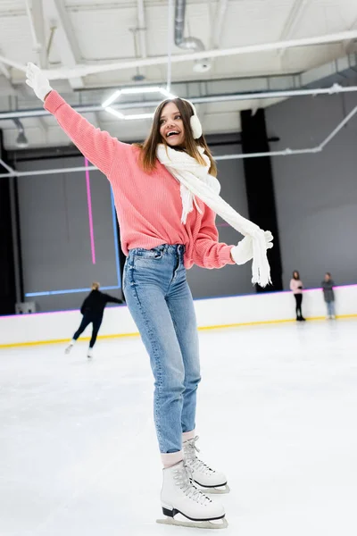 Pleine longueur de femme excitée dans des cache-oreilles blancs tenant écharpe tout en patinant avec la main tendue sur la patinoire — Photo de stock