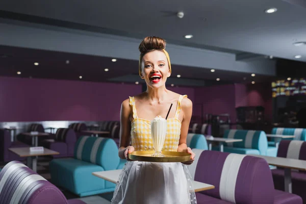 Alegre pin up camarera en vestido sosteniendo bandeja con batido en la cafetería - foto de stock