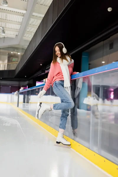 Comprimento total de feliz jovem mulher em roupa de inverno olhando para patins de gelo na pista de gelo — Fotografia de Stock
