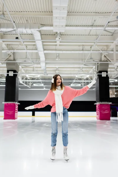 Longitud completa de la mujer sorprendida en bufanda blanca, suéter y orejeras patinaje en pista de hielo - foto de stock
