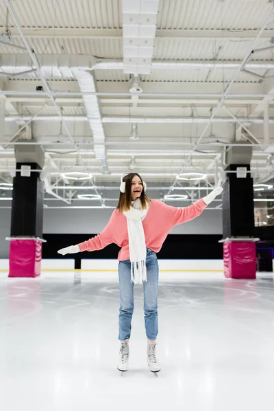 Piena lunghezza di donna stupita in sciarpa bianca, maglione e paraorecchie pattinaggio su pista di pattinaggio — Foto stock