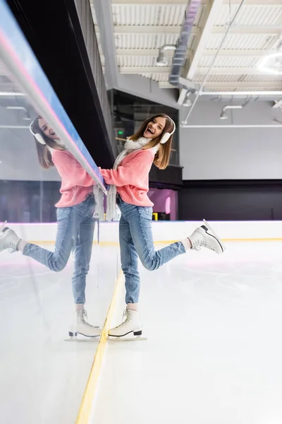 Longitud completa de la mujer sorprendida en bufanda blanca, suéter y orejeras posando en pista de hielo - foto de stock