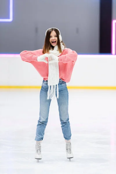 Comprimento total de mulher alegre em cachecol branco e abafadores de ouvido mostrando coração com as mãos enquanto patinava na pista de gelo — Fotografia de Stock
