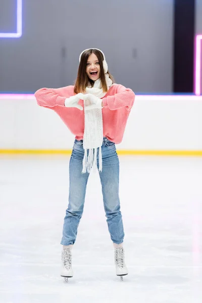 Pleine longueur de femme gaie en écharpe blanche et cache-oreilles montrant le cœur avec les mains tout en patinant sur la patinoire — Photo de stock