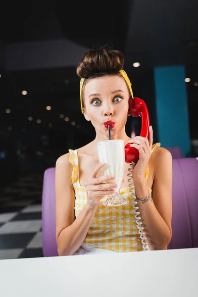 Шокована жінка розмовляє на ретро телефоні та п'є молочний коктейль — стокове фото