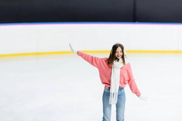 Mulher sorridente em regalos de orelha branca, cachecol e suéter patinação com a mão estendida na pista de gelo — Fotografia de Stock