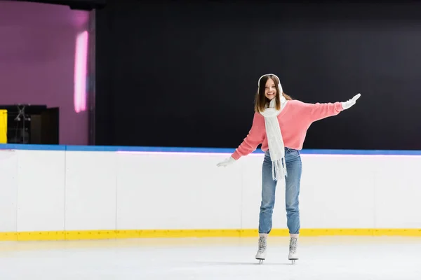 Longitud completa de la mujer sonriente en orejeras y traje de invierno patinaje en pista de hielo - foto de stock