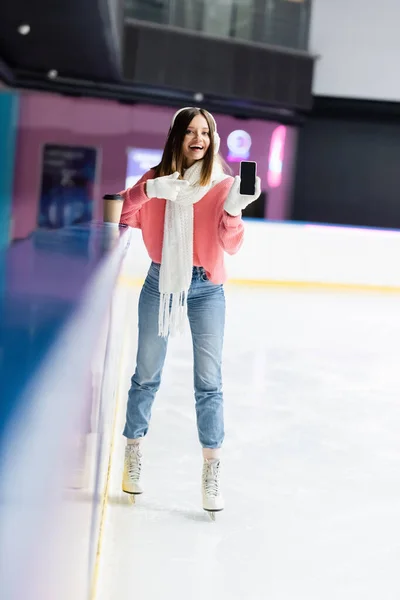 Mulher feliz em regalos de orelha branca e suéter rosa apontando com o dedo para o celular com tela em branco na pista de gelo — Fotografia de Stock