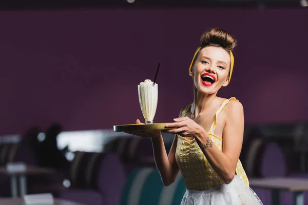 Asombrado pin up camarera en vestido celebración bandeja con batido en la cafetería - foto de stock