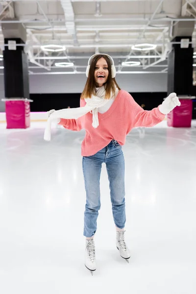 Comprimento total da mulher feliz em regalos auriculares e roupas de inverno patinação e segurando cachecol na pista de gelo — Fotografia de Stock