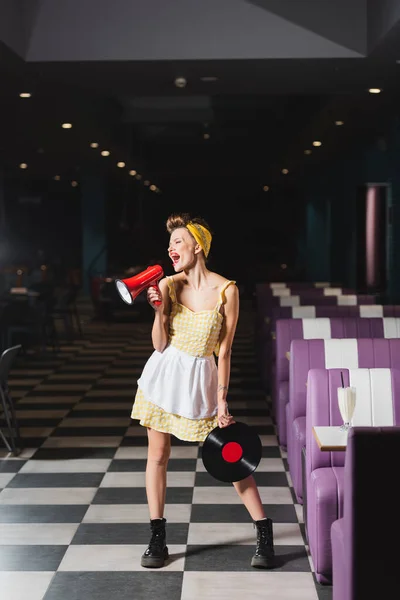 Pin up woman screaming in megaphone and holding vinyl disc in cafe — Stock Photo