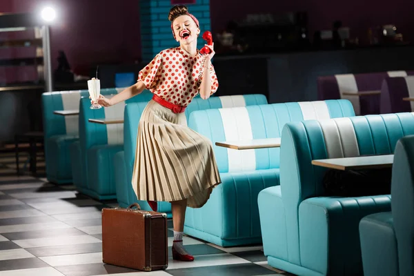 Amazed pin up woman holding milkshake cocktail and retro telephone near suitcase in cafe — Stock Photo