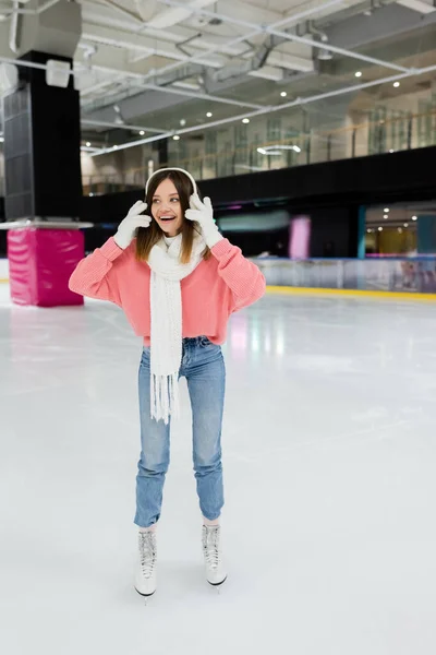 Full length of cheerful young woman in white ear muffs and winter outfit skating on frozen ice rink — Stock Photo
