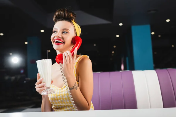 Happy pin up woman holding tasty milkshake and talking on retro telephone in cafe — Stock Photo