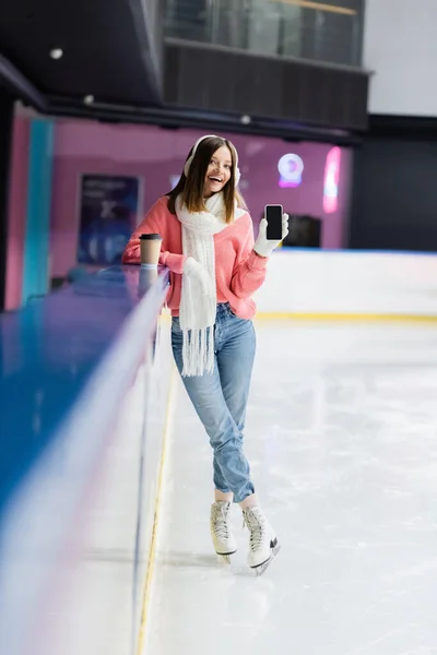 Full length of happy woman in ear muffs and gloves holding smartphone with blank screen on ice rink — Stock Photo
