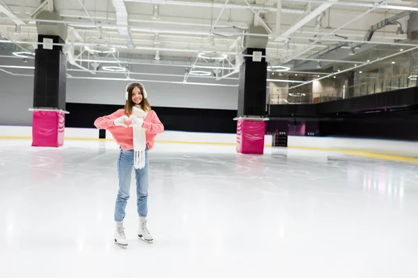 Full length of happy woman in white scarf and ear muffs showing heart with hands while skating on ice rink — Stock Photo