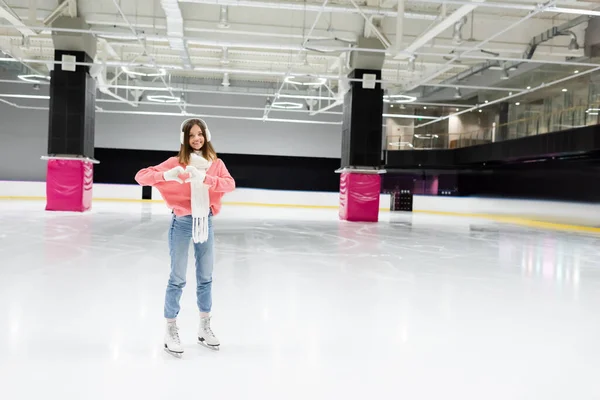 Pleine longueur de femme heureuse en écharpe blanche et cache-oreilles montrant le cœur avec les mains tout en patinant sur la patinoire — Photo de stock