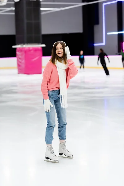 Comprimento total da mulher feliz em suéter, cachecol branco e abafadores de ouvido patinando na pista de gelo — Fotografia de Stock