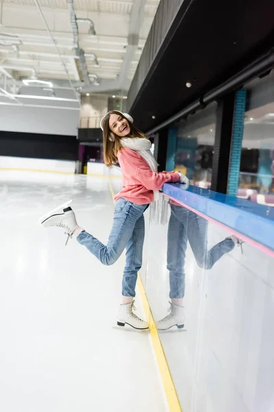 Full length of happy woman in sweater, white scarf and ear muffs posing on ice rink — Stock Photo