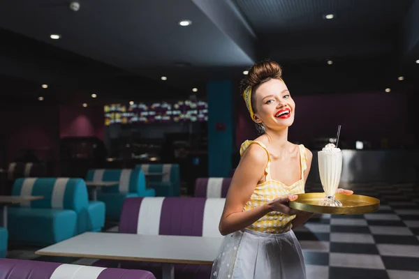 Alegre camarera en pin up vestido bandeja de celebración con batido en la cafetería - foto de stock