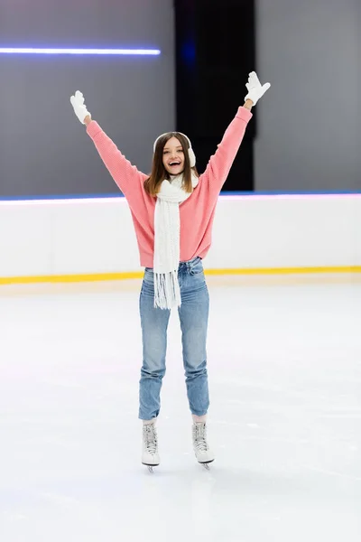 In voller Länge glückliche Frau in Strickpullover, Ohrenschützer und Winteroutfit beim Schlittschuhlaufen mit erhobenen Händen auf der Eisbahn — Stockfoto