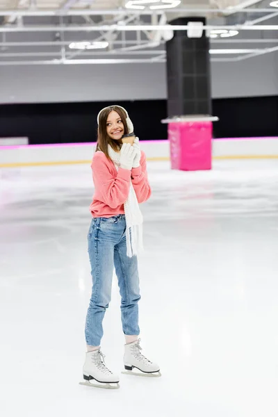 Full length of happy woman in knitted sweater, ear muffs and winter outfit skating with paper cup on ice rink — Stock Photo