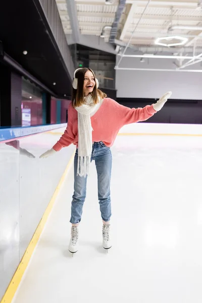 Longitud completa de la mujer feliz en suéter de punto, orejeras y patinaje traje de invierno mientras agita la mano en la pista de hielo - foto de stock