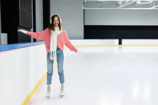 Full length of happy woman in knitted pink sweater, ear muffs and winter outfit skating on ice rink — Stock Photo