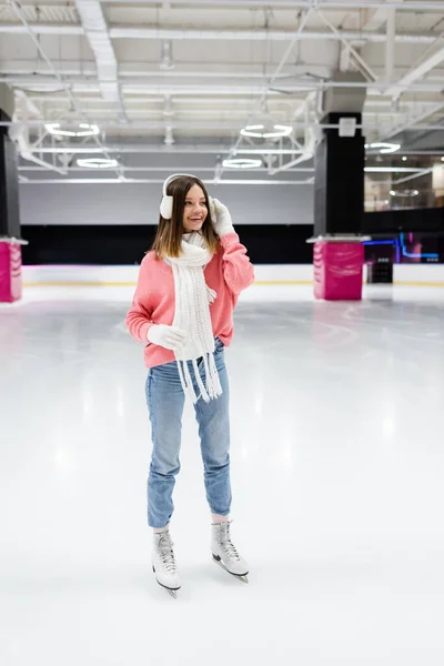 Comprimento total da mulher feliz em camisola rosa de malha, abafadores de orelha e roupas de inverno patinação na pista de gelo congelada — Fotografia de Stock